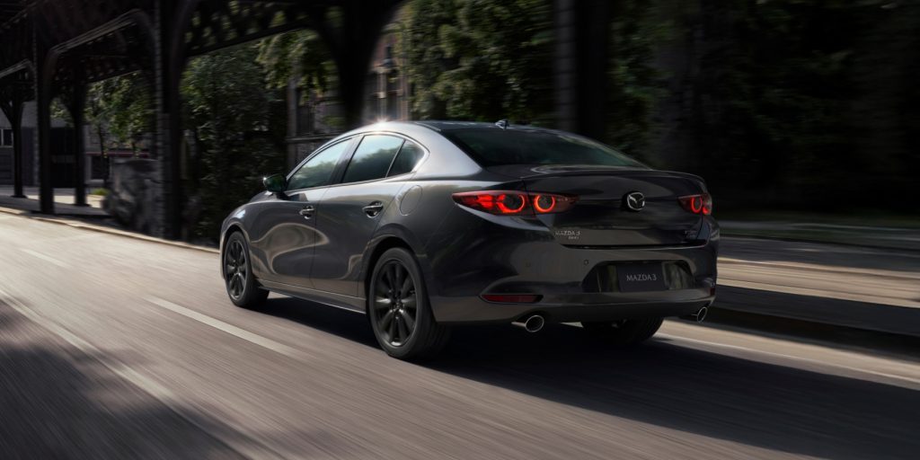 Rear view of a gray 2026 Mazda3 sedan driving on a city street with sleek LED taillights and a sporty design.