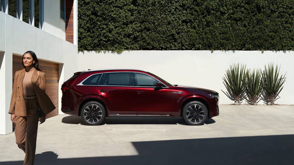 Side view of a red 2026 Mazda CX-90 parked outside a modern home with a person walking nearby.