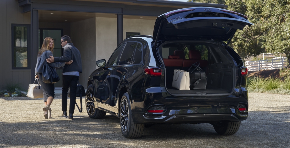 Rear view of a 2026 Mazda CX-70 with the liftgate open, showing spacious cargo area with luggage while a couple walks toward a house.